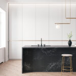 A modern kitchen features a black marble island with a sink and faucet. A wooden stool and pendant lights are nearby, while white cabinets and a large window brighten the room.