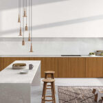 A modern kitchen features a long, white marble countertop with wooden barstools, pendant lights overhead, and a woven rug on the floor, creating a clean, minimalist atmosphere.