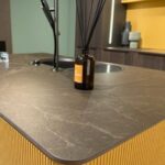 Caesarstone Porcelain 514 Emprada surface in Modern kitchen island with dark countertop, sleek faucet, reed diffuser, and wooden paneling.