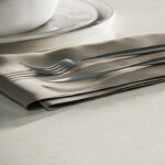 A fork and knife rest on a neatly folded beige napkin beside a white bowl and plate, set on a smooth, light-colored surface.
