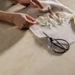 Close-up of Caesarstone 584 Opal Taj countertop surface featuring a person hand-embroidering a floral pattern on linen cloth with vintage scissors.