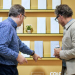 Two men examine a wall display with various paper samples and a small potted plant.
