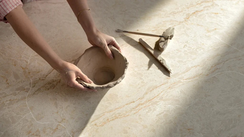 Caesarstone IcON 8251 Taj Whisper Hands shaping a clay bowl, with sculpting tools nearby on a marble surface.