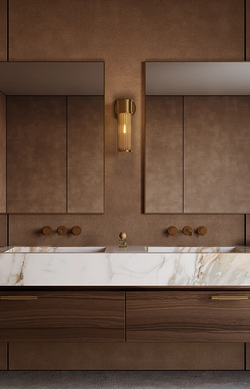 Double vanity with marble countertop, two mirrors, bronze fixtures, warm lighting, and brown textured walls.